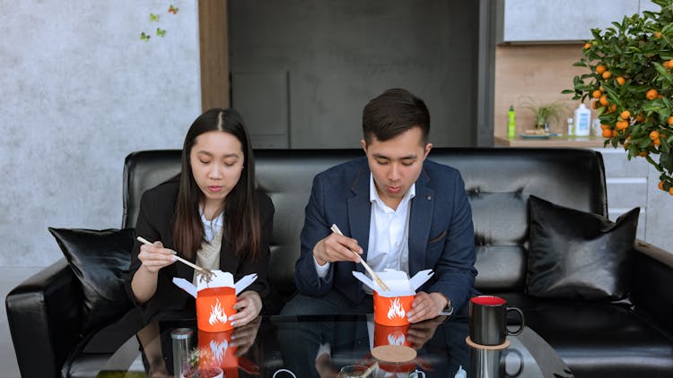 Man And Woman Sitting On Black Couch  Eating Food On A Takeout Box With Chopsticks