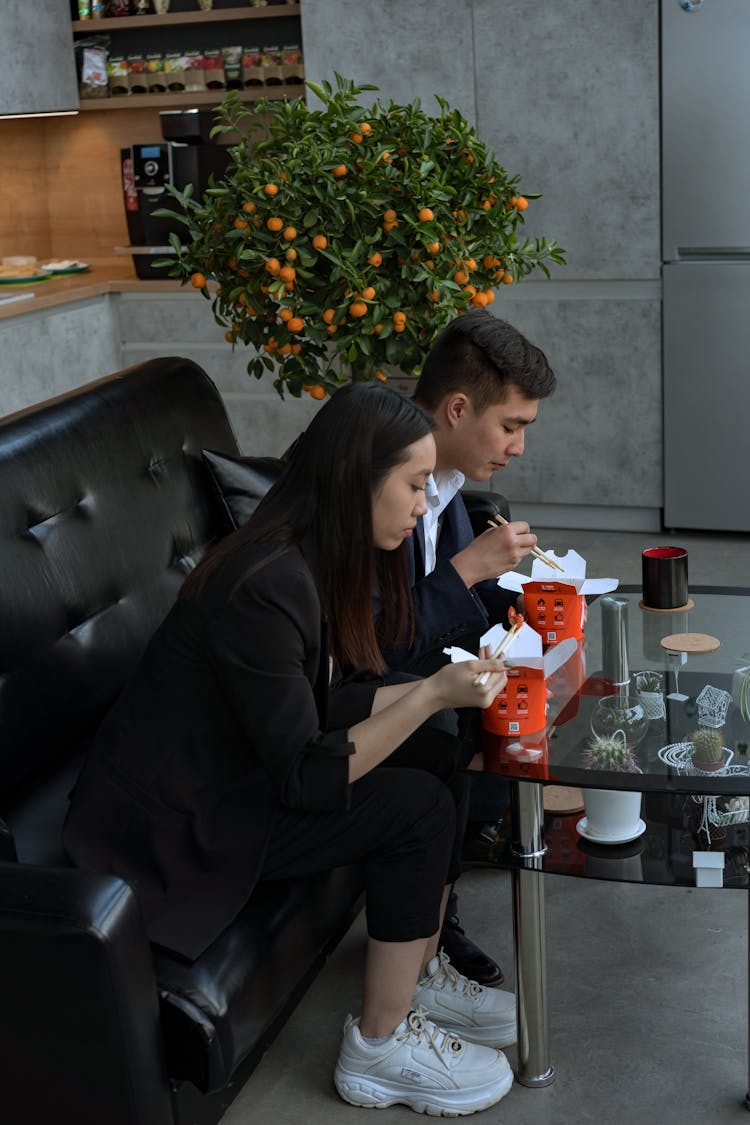 Man And Woman Sitting On Leather Sofa Eating On Takeout Boxes Using Chopsticks