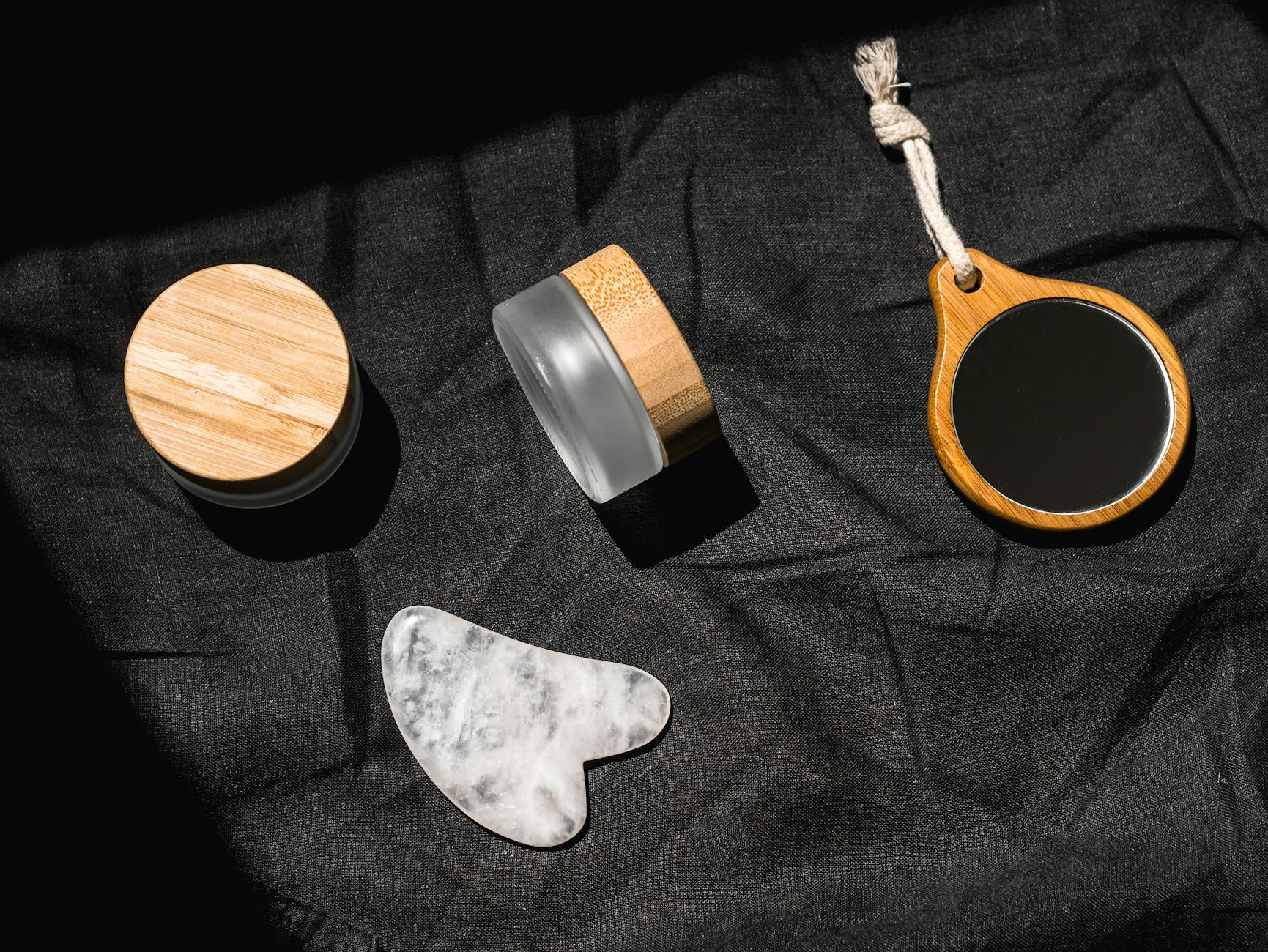 A flat lay of eco-friendly beauty tools on black fabric, including a wooden mirror and bamboo jar.