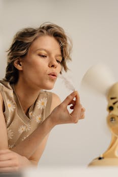 Young woman playfully blowing a feather in front of an electric fan indoors.