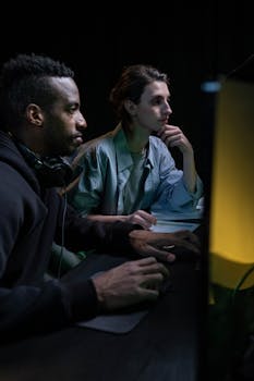 Two colleagues collaborating in a dimly lit office setting, immersed in computer work.