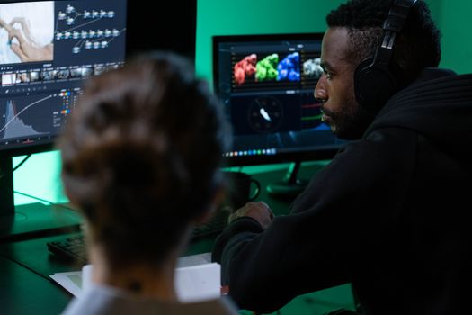 A man wearing headphones works on video editing with dual monitors in a dimly lit room.