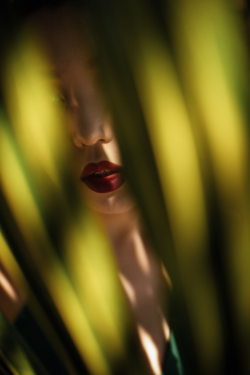 Enigmatic portrait of a woman partially hidden by sunlit palm leaves, rich red lips in focus.