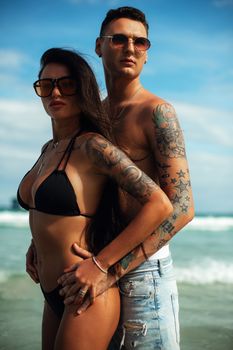 A fashionable couple in swimwear and sunglasses posing confidently on a sunny beach.