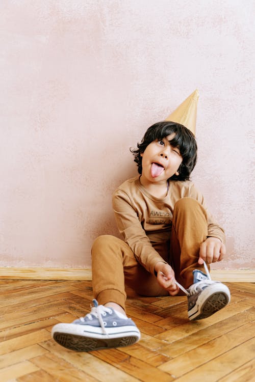 A Young Boy in Brown Sweater and Pants Sitting on a Wooden Floor while Wearing a Party Hat