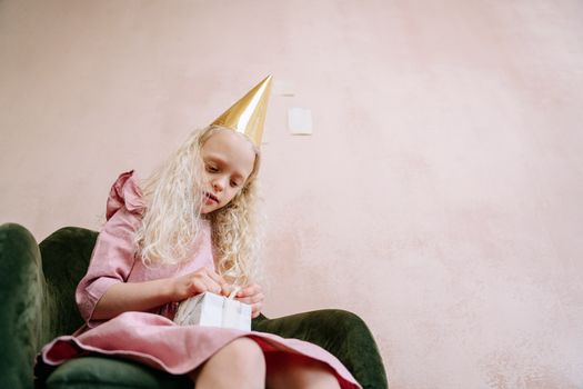 A young girl in a pink dress and party hat opening a gift indoors.