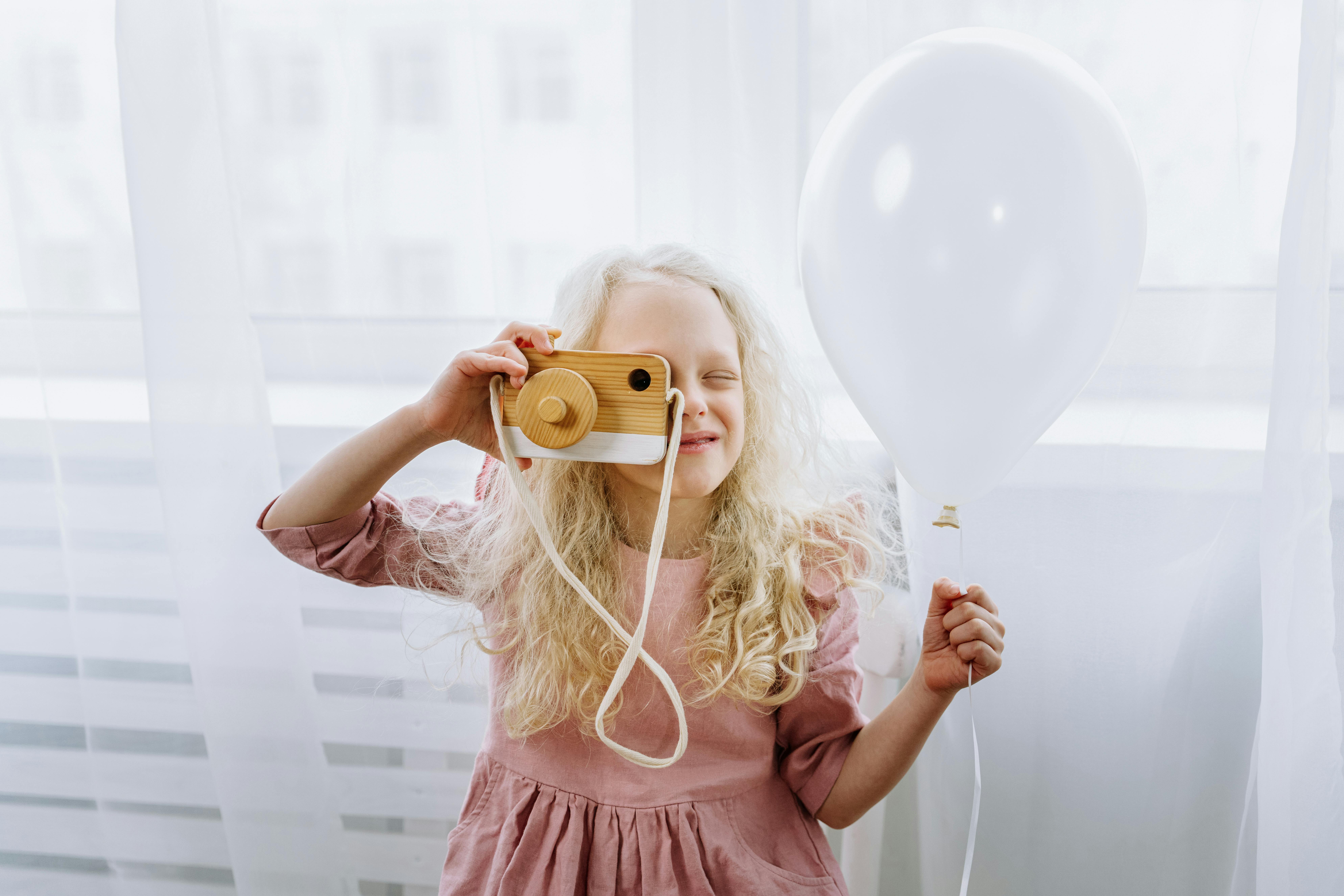 A Girl in Pink Dress Holding a Wooden Toy Camera and White Balloon ...