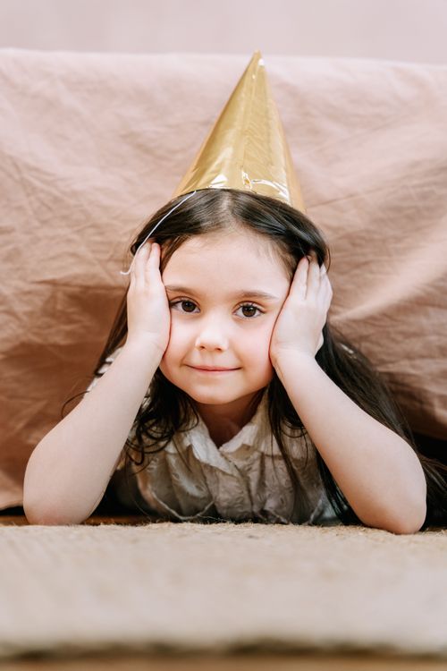 Free A cheerful child lying down, wearing a gold party hat indoors. Perfect for festive and joyful themes. Stock Photo