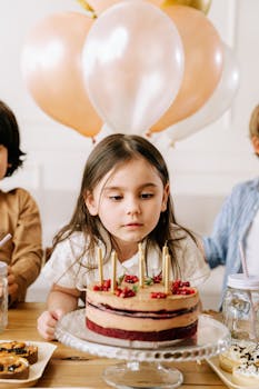 A young girl at a birthday party prepares to blow out candles on a cake surrounded by balloons.