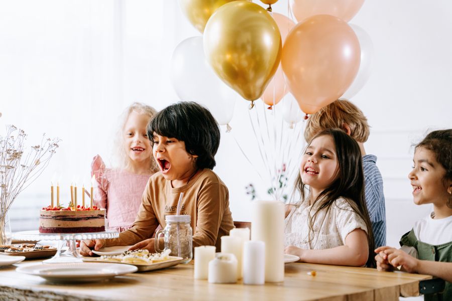 Children gathered around a birthday cake with balloons in the background.