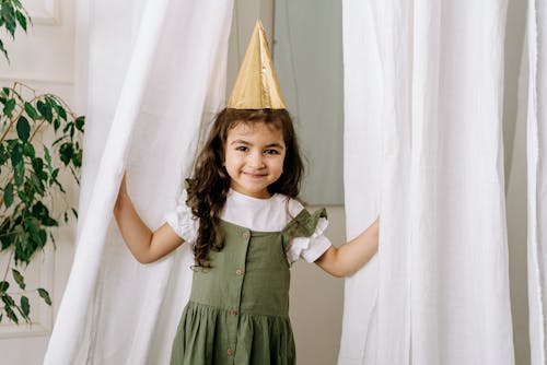Girl Wearing a Party Hat Standing Near White Curtains