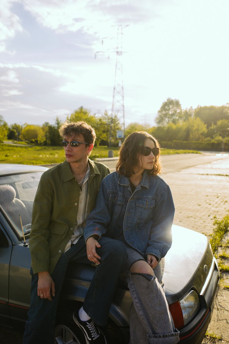 A Couple In Green And Denim Jacket Sitting On The Car Hood While Wearing Sunglasses