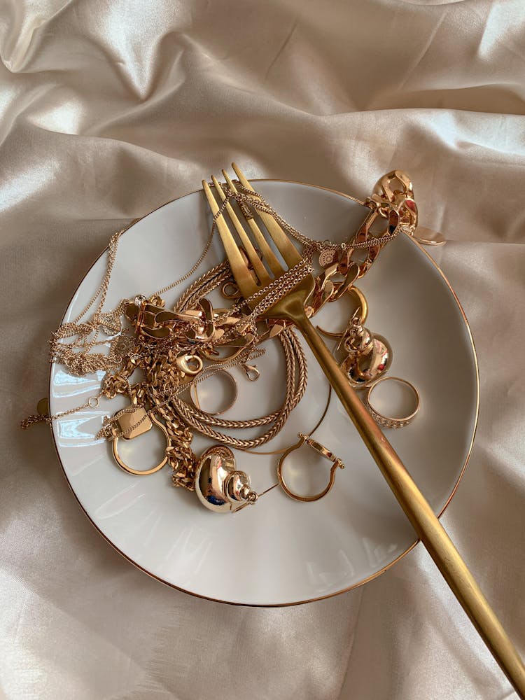 Golden Jewelry And Golden Fork On White Saucer On Beige Silk