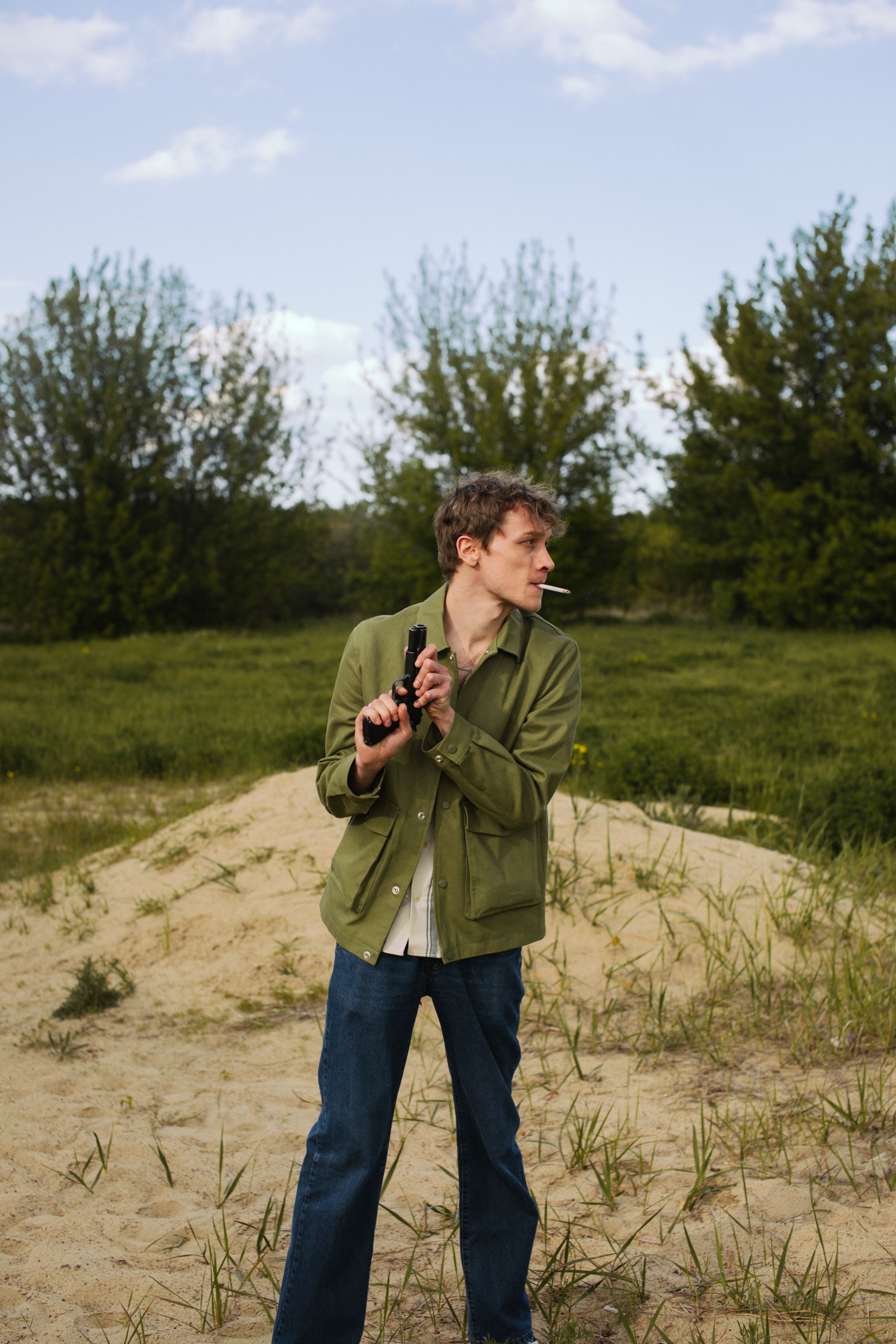 A man stands outdoors holding a gun and smoking a cigarette, surrounded by trees and sand.