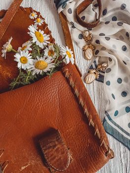 Charming arrangement of a leather bag with daisies and jewelry on a wooden surface.
