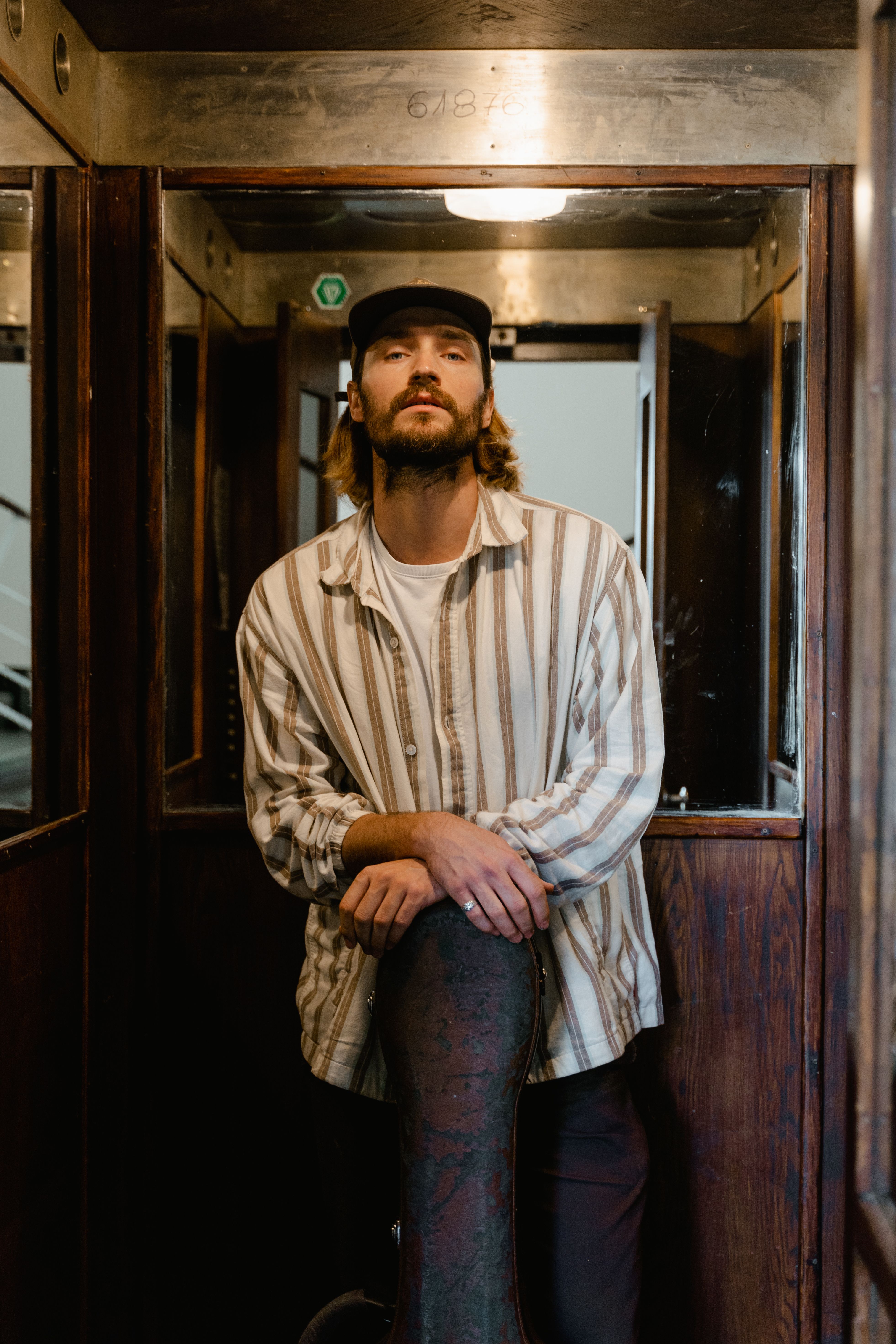 Musician standing with guitar case in a vintage train compartment, indoors.