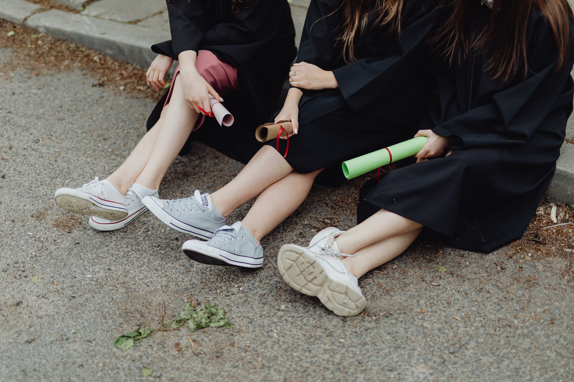 Three graduates in gowns sit on a roadside holding diplomas, legs crossed.