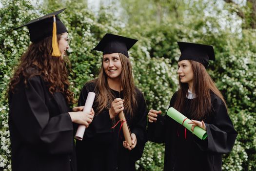 Three young women in caps and gowns celebrate their graduation with diplomas outdoors.