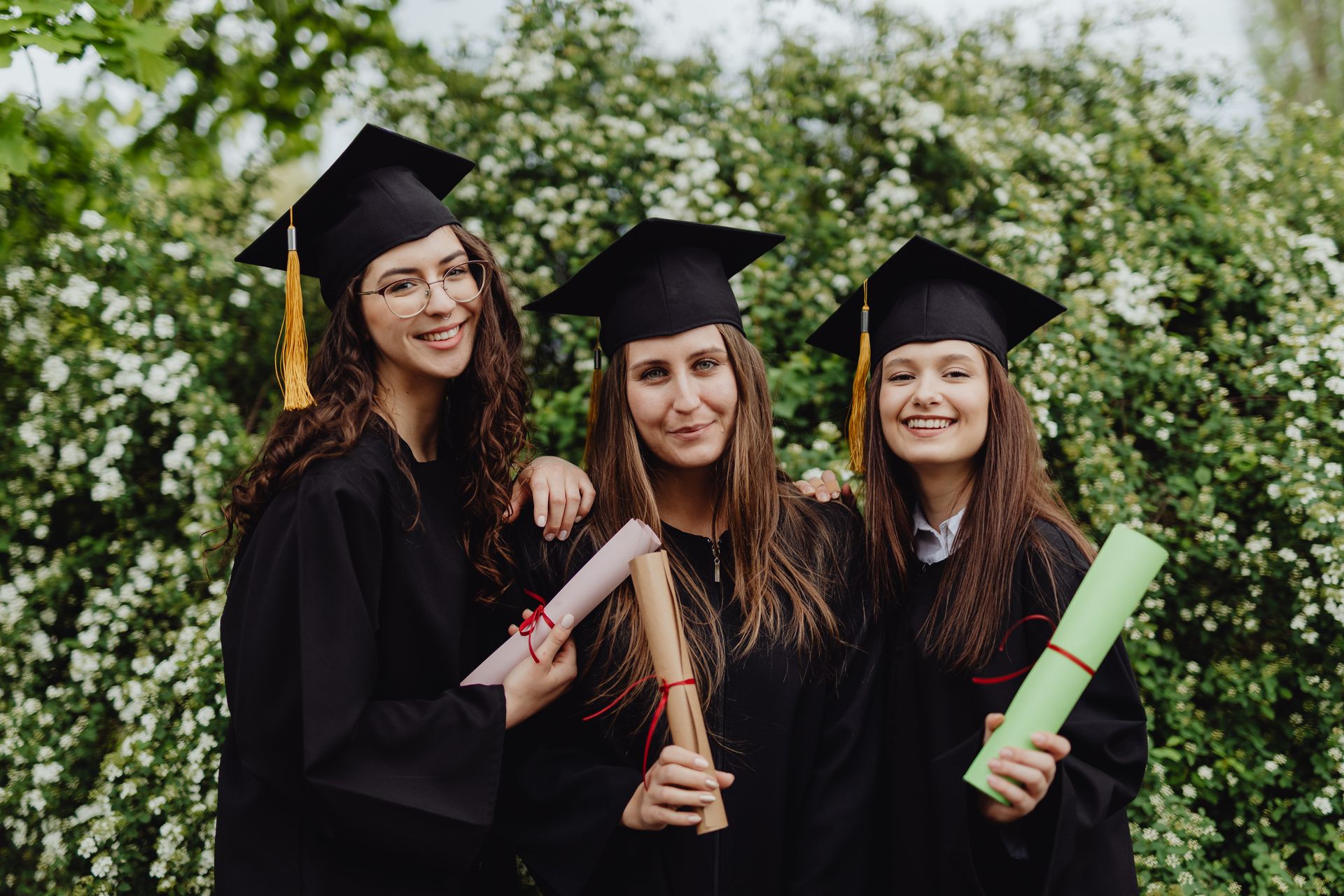 Three young female graduates in caps and gowns celebrating with diplomas outdoors.