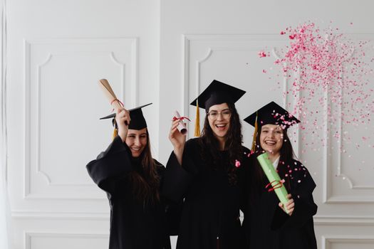 Three young female graduates celebrating indoors with diplomas and confetti.