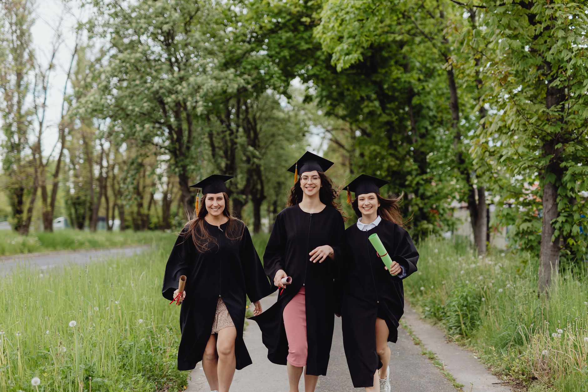 graduation party themes egypt — Three female graduates in academic robes, celebrating outdoors in a lush green environment.