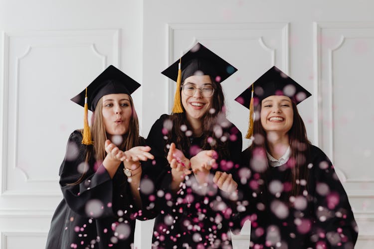 Women In Graduation Gowns And Caps Blowing Confetti