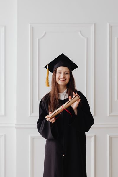 Happy young woman in graduation gown holding diploma indoors.