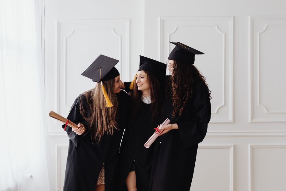 Three young women in graduation gowns and caps celebrate together indoors with diplomas in hand.