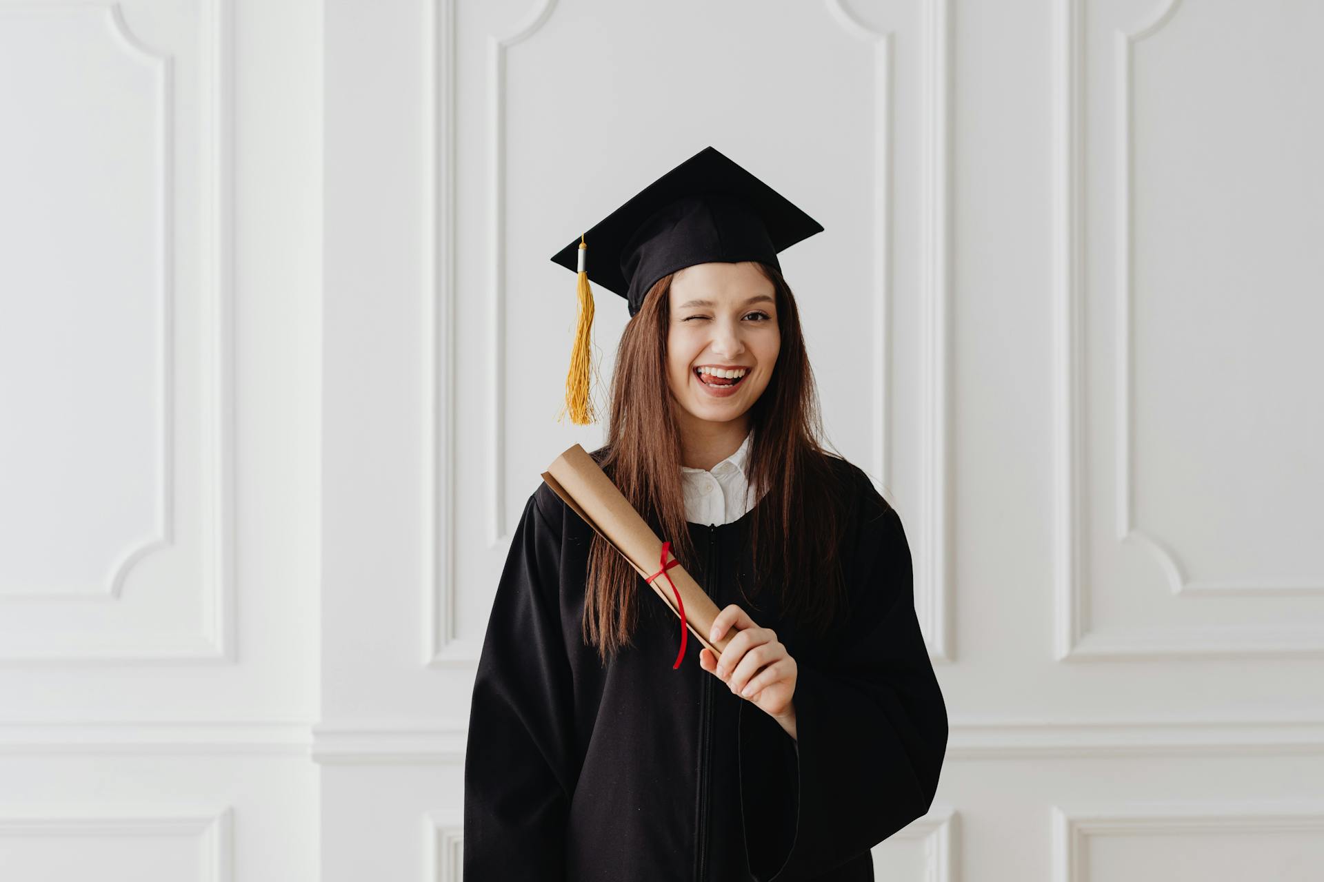 Happy graduate in cap and gown holding diploma smiling indoors, celebrating academic achievement.