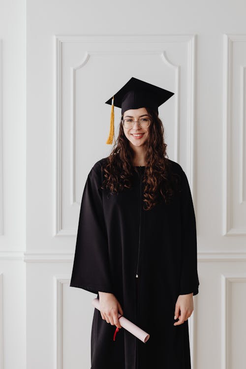 Free Woman in Black Academic Dress Standing Near White Wall Stock Photo