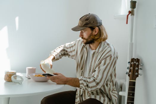 Man pouring milk into cereal while using smartphone at breakfast table.