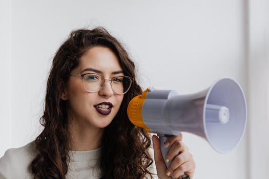 Confident woman with eyeglasses speaking into a megaphone in a bright room.