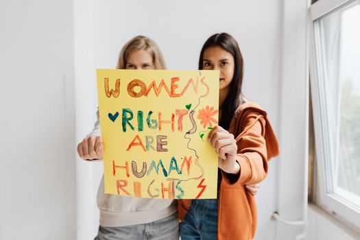 Two women confidently hold a vibrant protest sign advocating women's rights indoors.