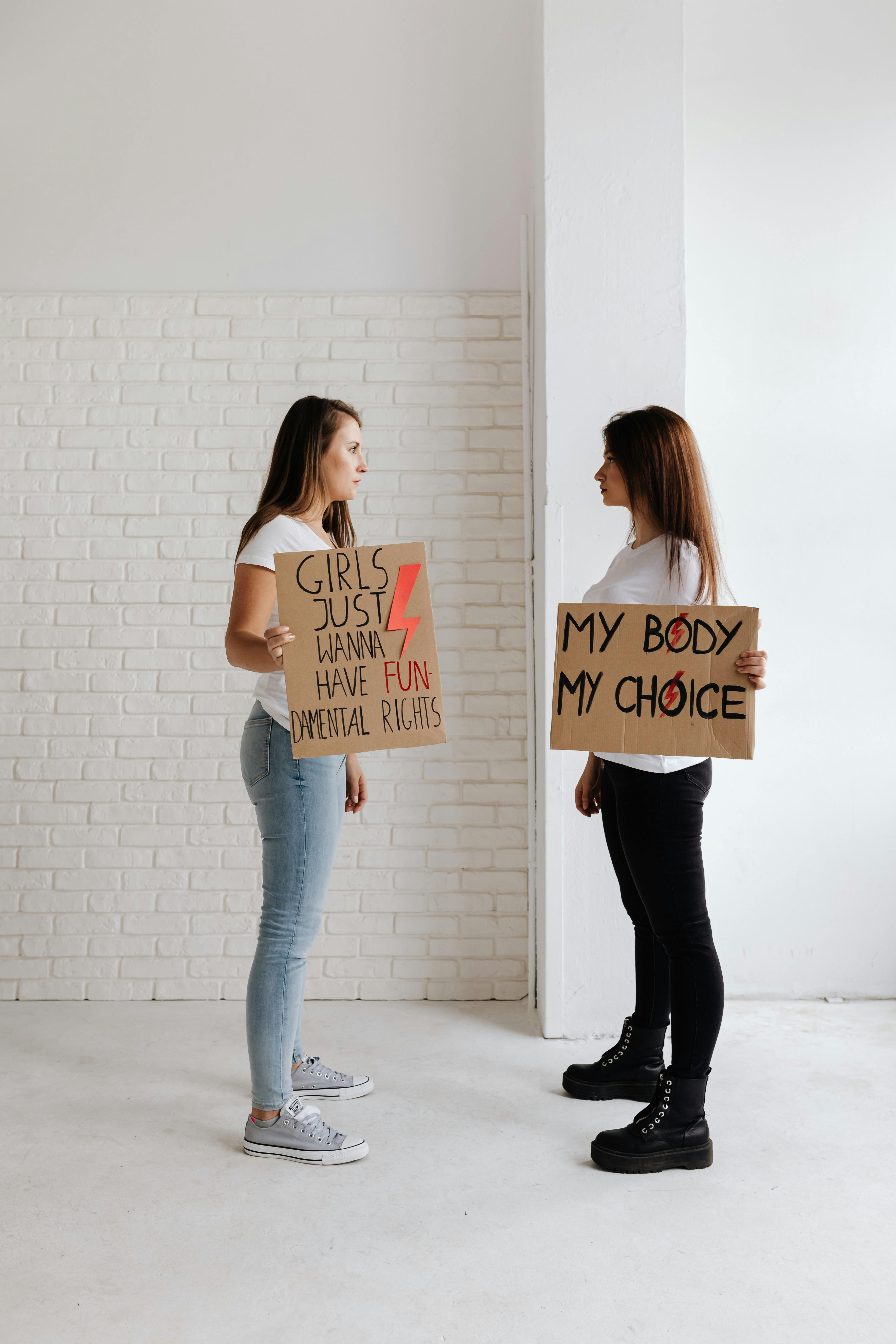 Two women standing in a white room holding signs advocating for women's rights.