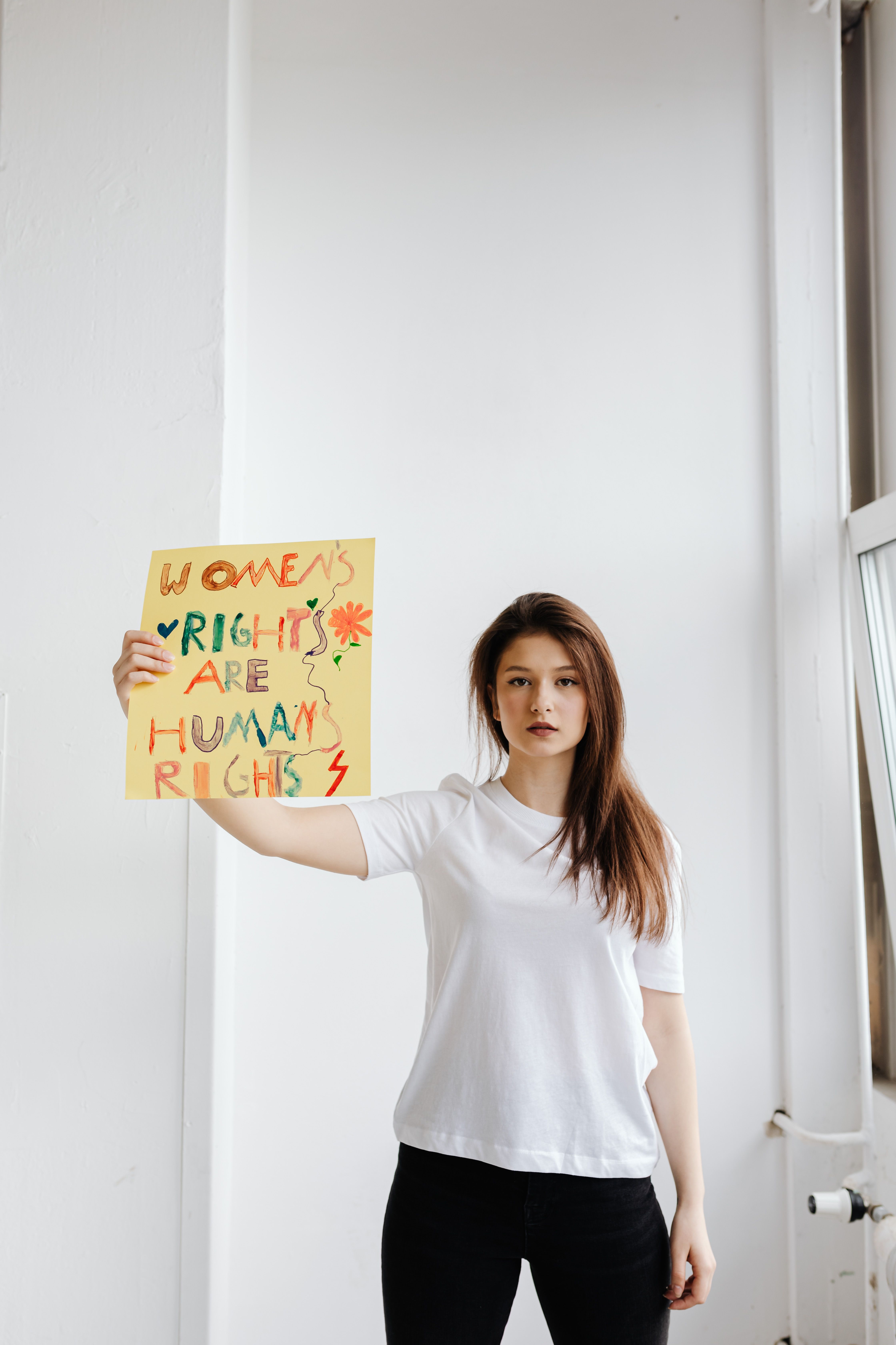 A young woman stands indoors holding a vibrant women's rights protest sign.