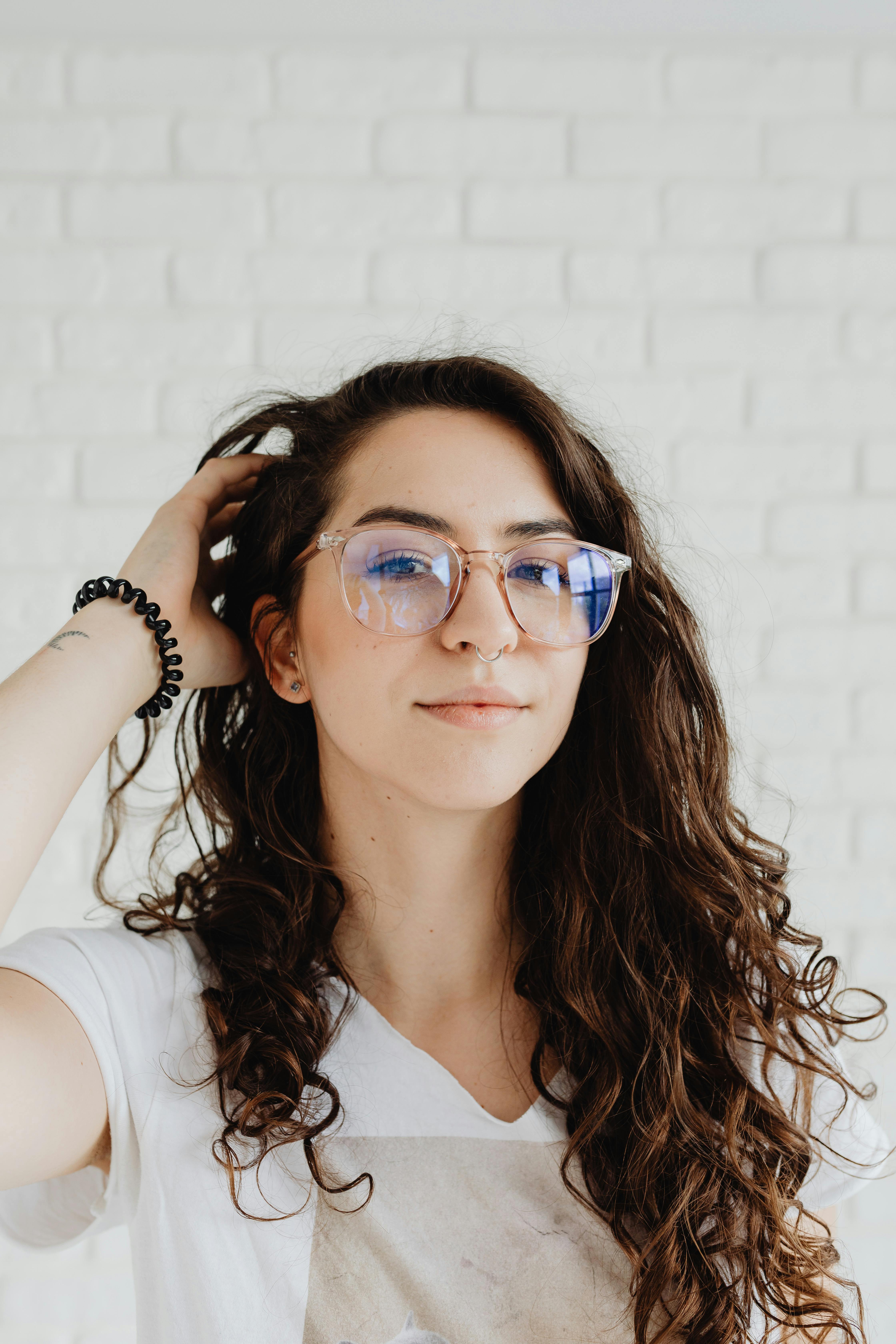 一位自信的年轻女子戴着眼镜，穿着白色衬衫，在室内砖墙前摆姿势