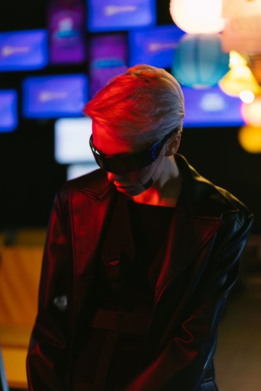 Stylish woman in cyberpunk sunglasses and leather jacket illuminated by neon lights, embodying futuristic fashion.