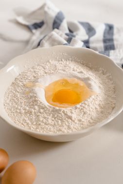 Tigela cerâmica com pó branco e ovo ao lado — ingredientes de panificação sobre superfície clara