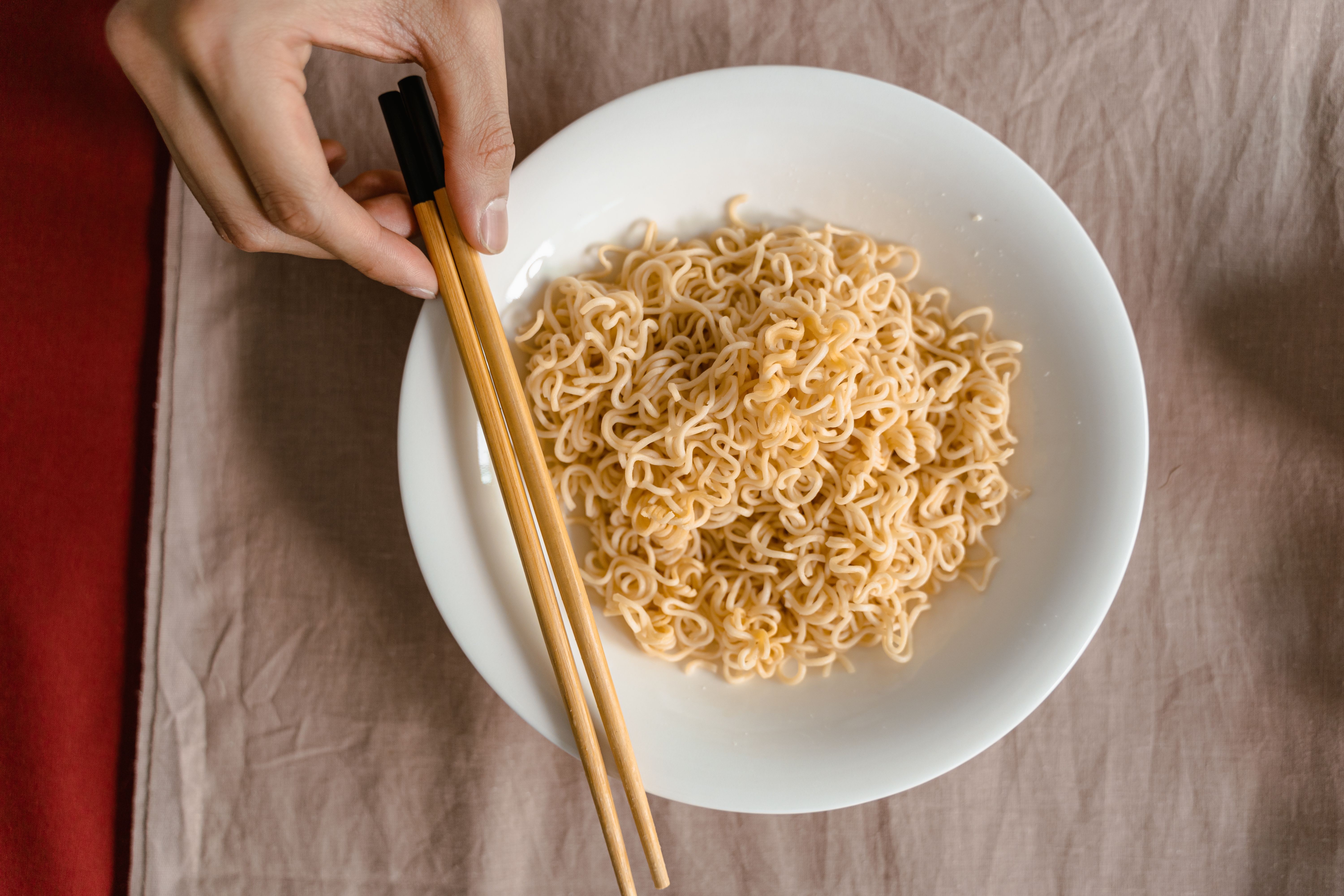 Close-up of a hand using chopsticks to pick noodles from a white ceramic bowl on a table.