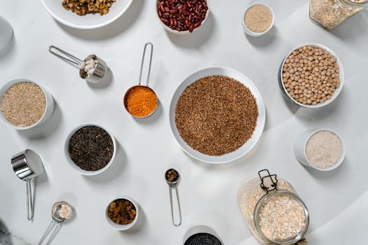 Top view of various grains, legumes, and spices in bowls and jars on a white table.