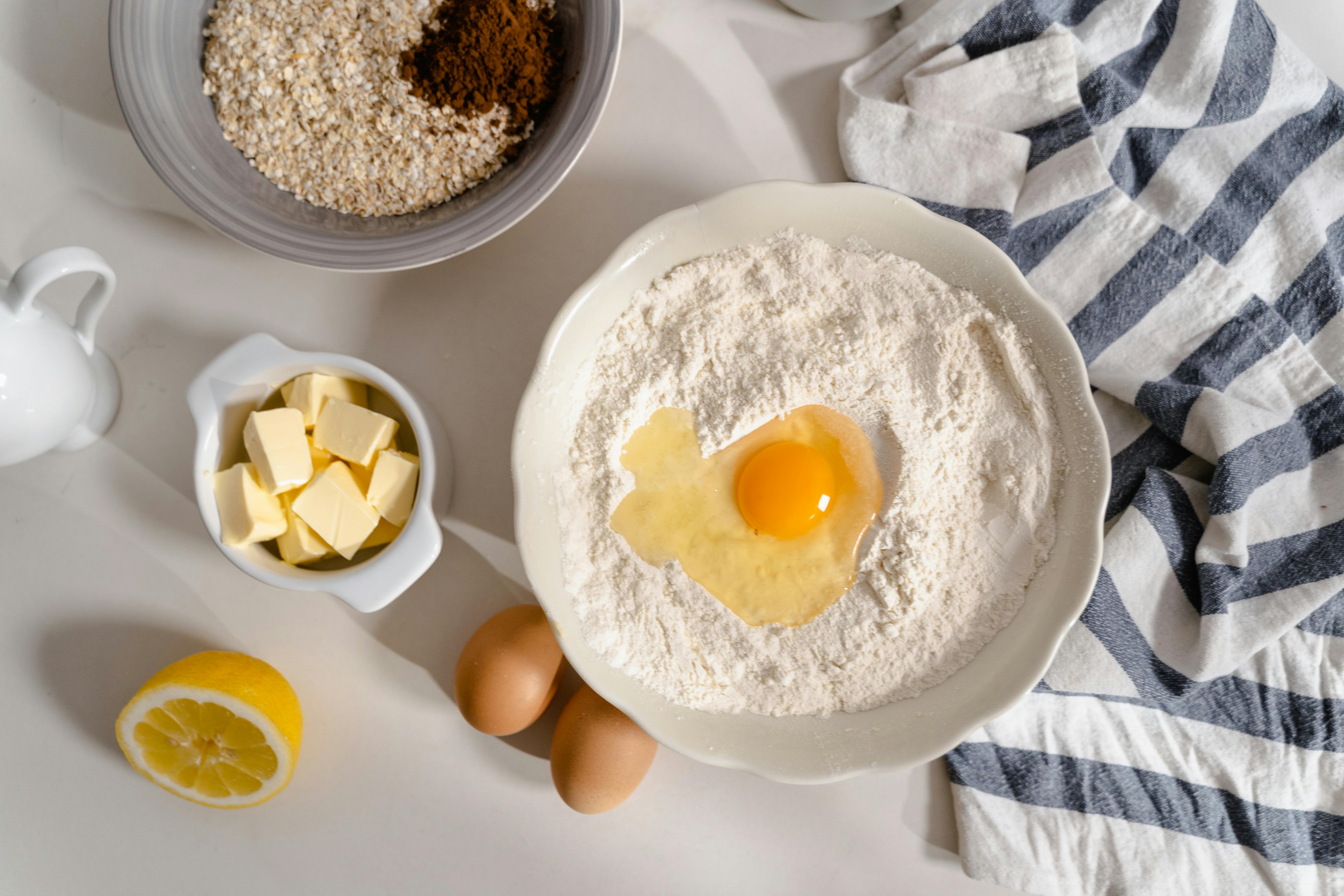 Flat lay photo of baking ingredients including flour, butter, and eggs on a white surface.