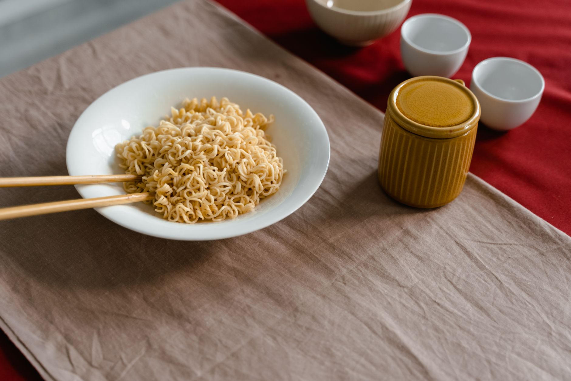 A plate of cooked noodles with chopsticks, ceramic mugs, and cups on a red and beige tablecloth.