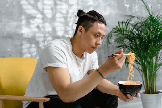 Asian man in white shirt enjoys a bowl of ramen with chopsticks indoors.
