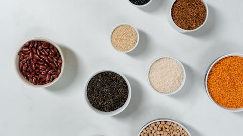 Flat lay of diverse grains and legumes in bowls on a marble surface, highlighting healthy ingredients.