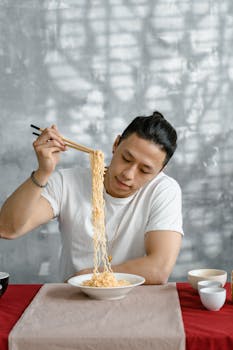 Asian man sitting at table, enjoying noodles with chopsticks indoors.