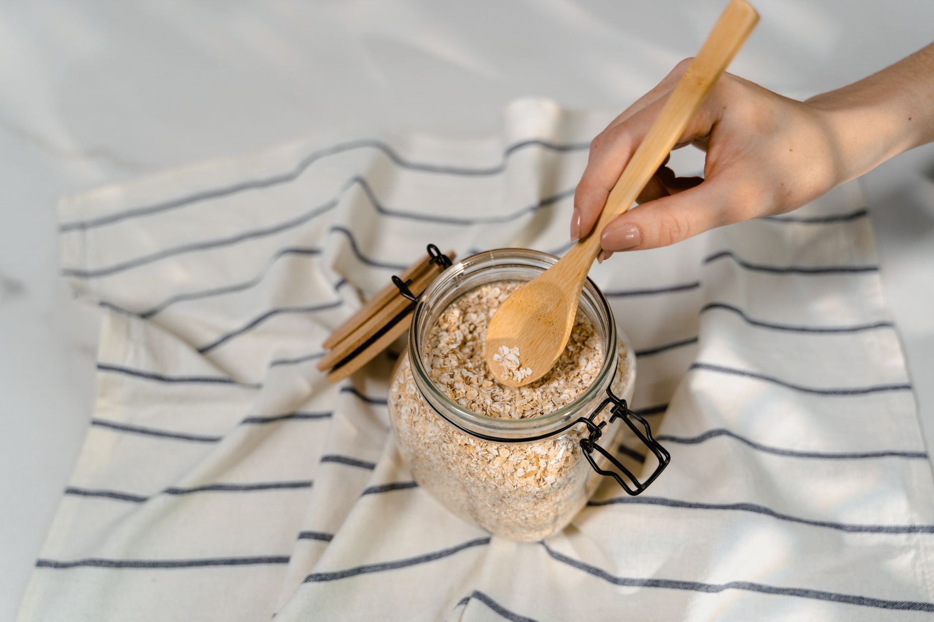 Close-up of a hand using a wooden spoon to scoop uncooked oatmeal from a glass jar on a striped cloth.