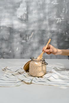 A hand stirring oats in a glass jar placed on a striped cloth, perfect for healthy breakfast themes.
