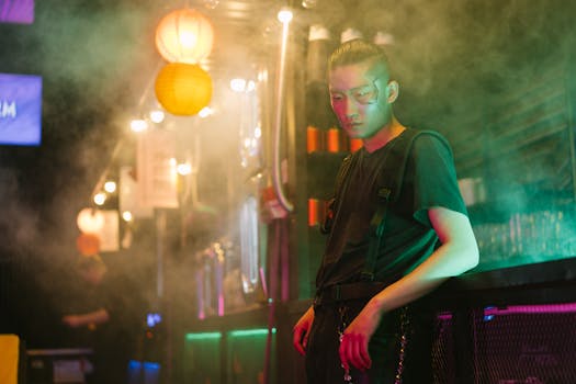 Asian man in cyberpunk setting with colorful lights and smoke, showcasing futuristic fashion.