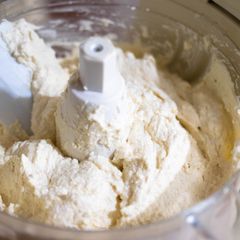 Close view into a food processor bowl of creamy batter around a white spindle, a white spatula at the side