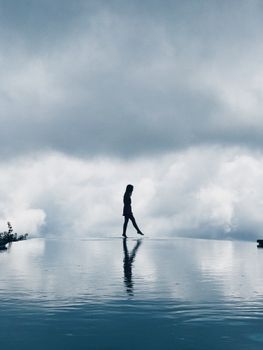 A dramatic silhouette of a woman walking by an infinity pool under a cloudy sky.